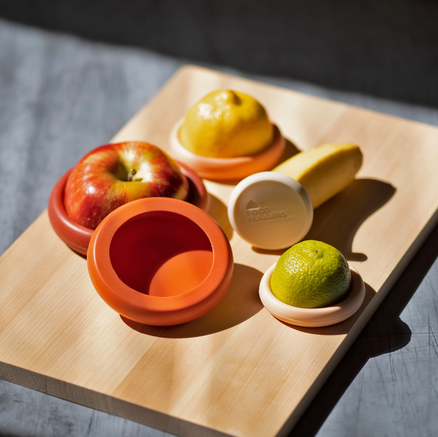 Apple, lemon, lime, and banana are partially covered with colorful Food Huggers by Food Huggers on a wooden cutting board in bright sunlight to help reduce food waste.