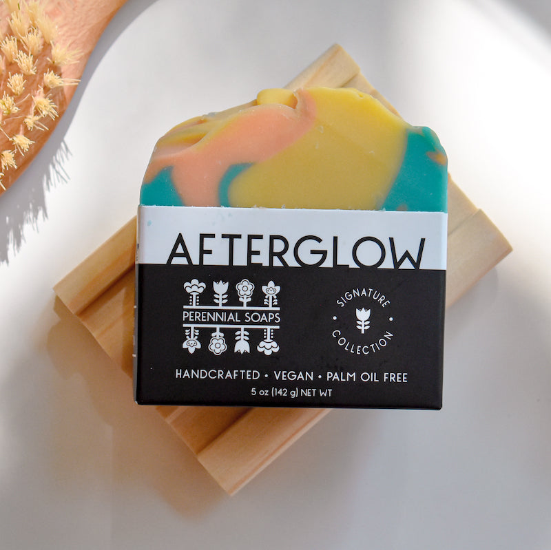 A vibrant Perennial Soaps Bar Soap with yellow, teal, and pink swirls rests on a wooden dish. The AFTERGLOW label notes its Wisconsin-made, vegan, palm oil free, and handcrafted.