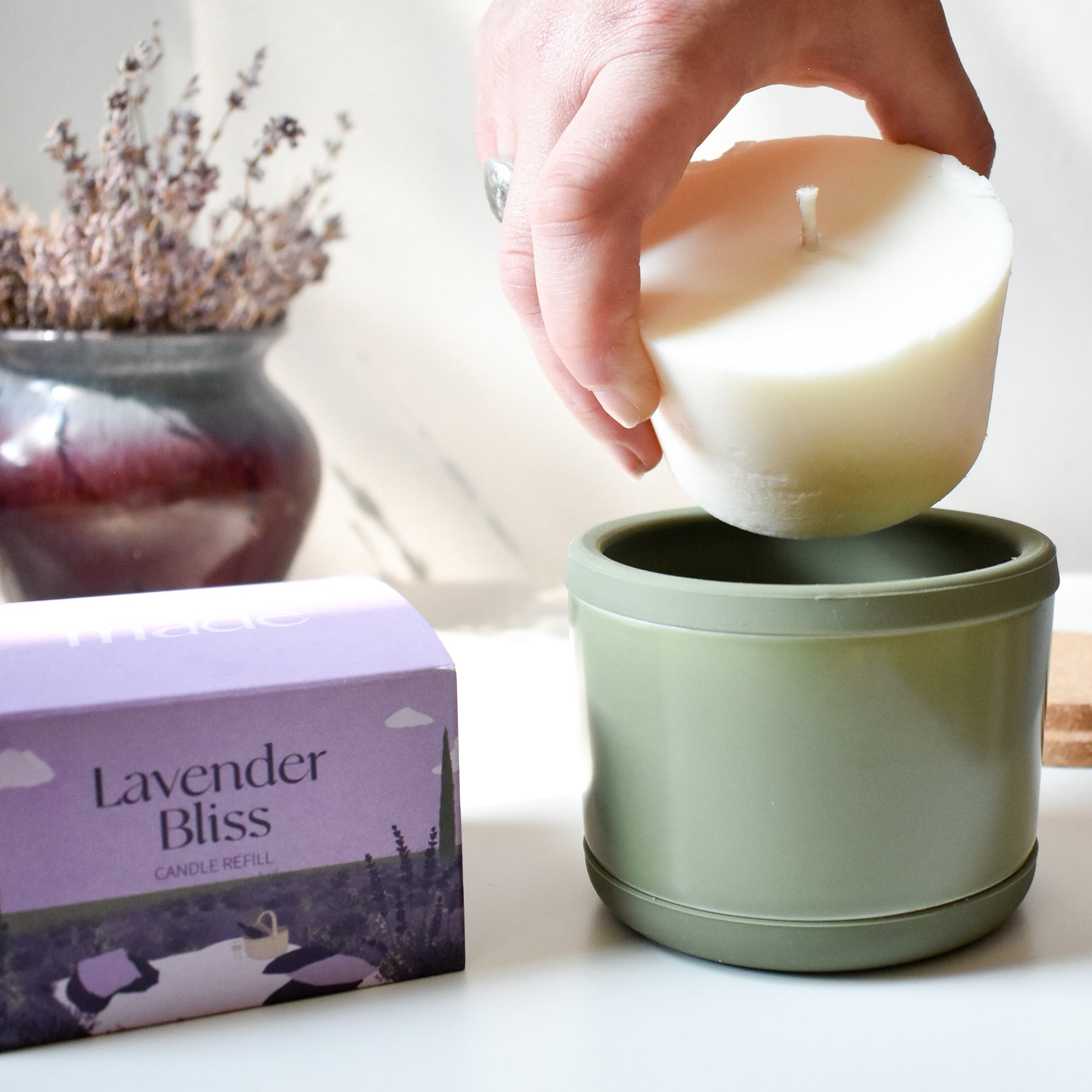 A hand places a white wax refill into an Arbor Made Refillable Candle Jar with a green silicone bottom. Beside it is a purple Arbor Made Lavender Bliss Candle Refill box, with a vase of dried lavender in the background.