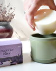 A hand places a white wax refill into an Arbor Made Refillable Candle Jar with a green silicone bottom. Beside it is a purple Arbor Made Lavender Bliss Candle Refill box, with a vase of dried lavender in the background.