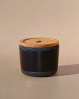 The Arbor Made Refillable Candle Jar by Arbor Made is a small, round black jar with a cork lid and silicone bottom. It sits on a beige surface, casting a soft shadow, while the neutral background highlights its elegant design.