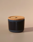 The Arbor Made Refillable Candle Jar by Arbor Made is a small, round black jar with a cork lid and silicone bottom. It sits on a beige surface, casting a soft shadow, while the neutral background highlights its elegant design.