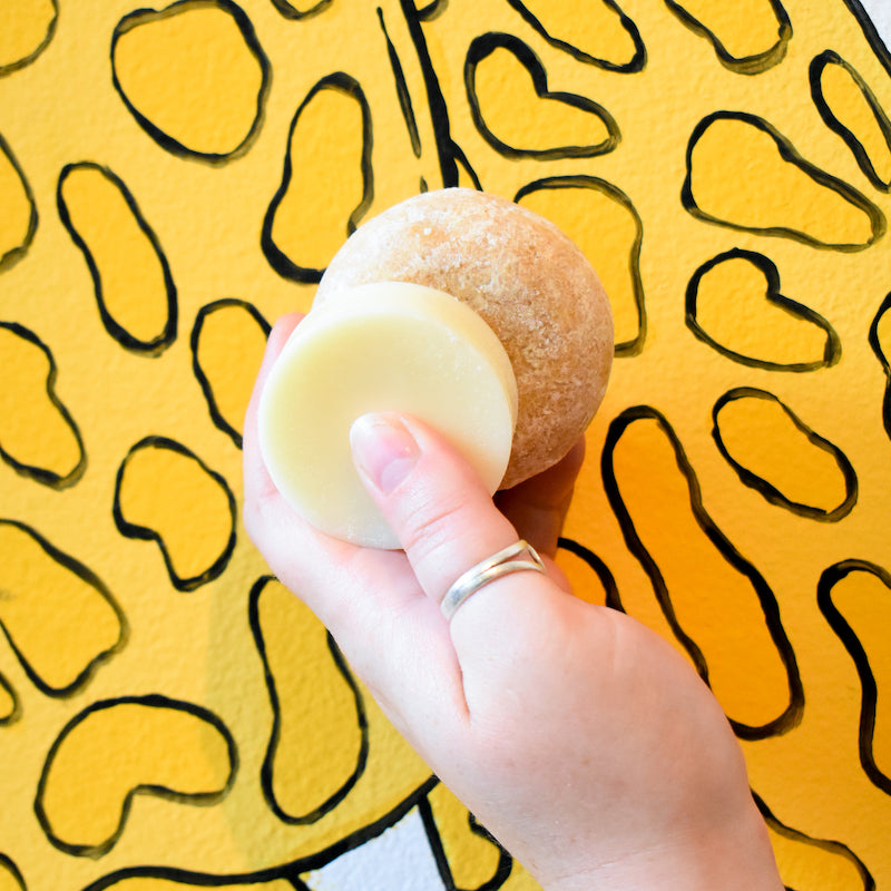 A hand holds two Notice Hair Co. Notice Shampoo & Conditioner Bars—one tan, one off-white—against a yellow background with black abstract shapes.