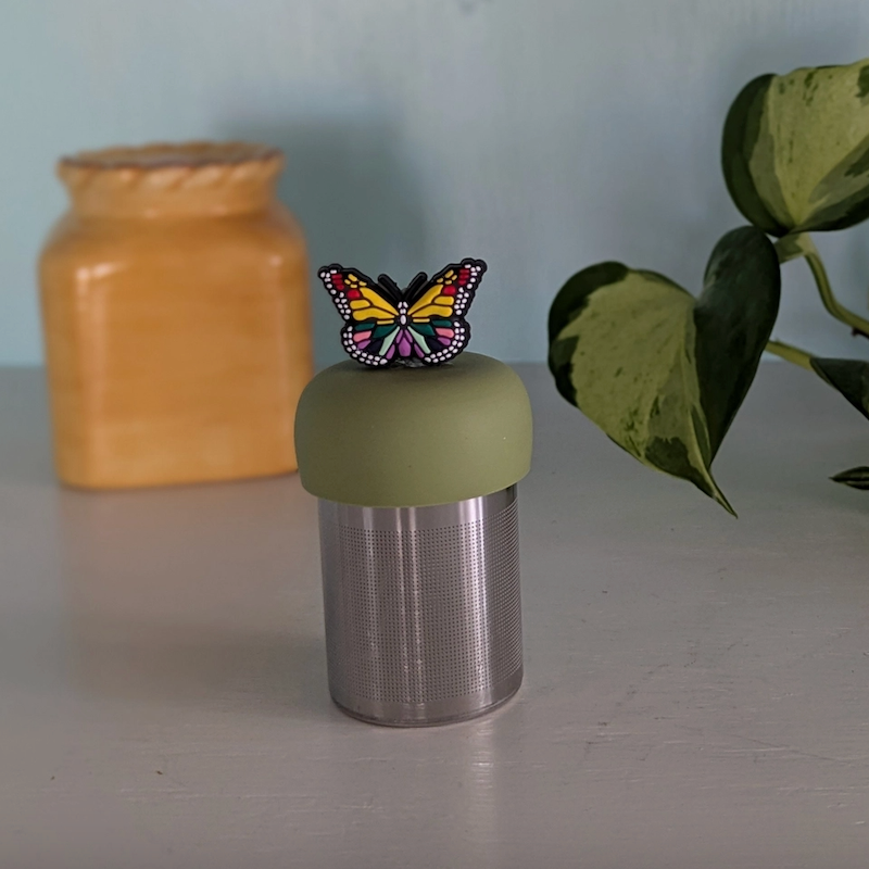 The Teany Tiny Kitchen Floating Tea Infuser features a metal container with a green lid and butterfly decoration. In the background, a yellow jar and leafy green plant sit on a light surface.