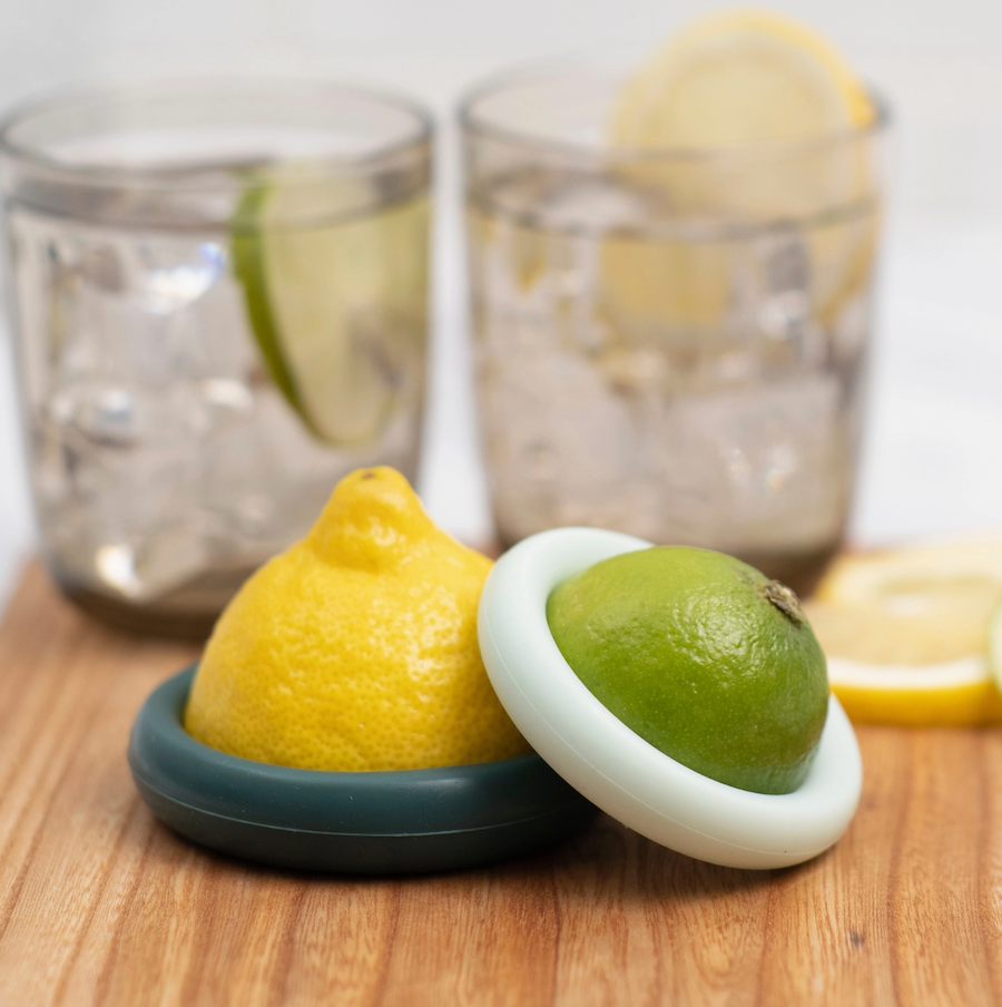 Halved lemon and lime sealed with Food Huggers by Food Huggers rest on a wooden surface in front of two glasses of iced water with citrus slices, illustrating an easy way to reduce food waste.
