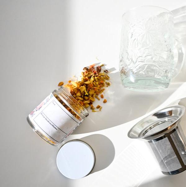 A glass jar of Glow Herbal Tea by Loose Leaf Tea Co. has spilled onto a white surface beside a clear textured glass mug and a metal tea infuser, with the jar’s lid set off to the side.