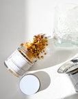 A glass jar of Glow Herbal Tea by Loose Leaf Tea Co. has spilled onto a white surface beside a clear textured glass mug and a metal tea infuser, with the jar’s lid set off to the side.