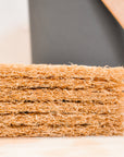 A stack of rectangular, rough-textured Bamboozle Hemp Compost Bin Filters is shown close-up with a blurred background and part of a black and wooden object, highlighting their natural fiber construction for odor control.