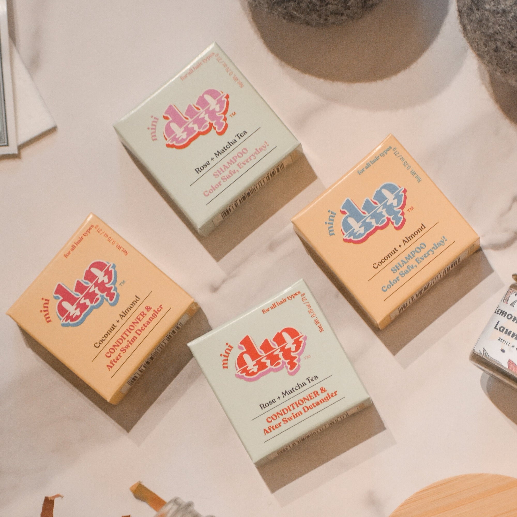 Four Dip Shampoo Bar Minis in pastel packaging (green, orange, yellow, beige) are arranged on a beige surface with other bath items nearby—compact and travel-friendly haircare from Dip.