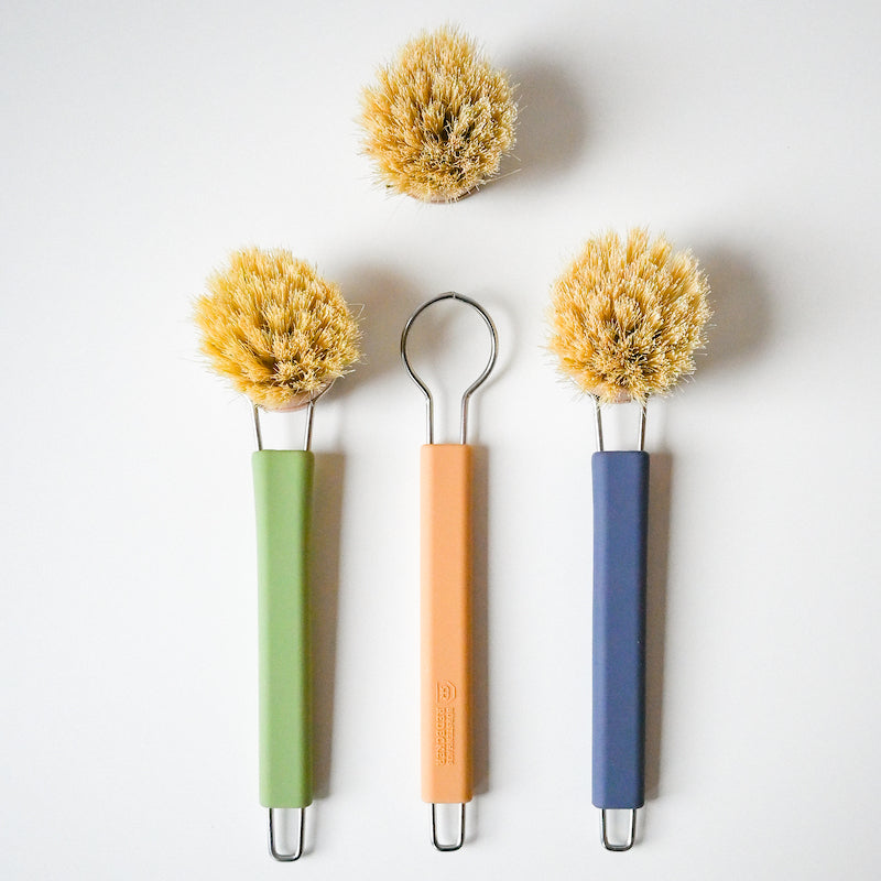 Three Redecker Dish Brushes With Removable Heads, each with a round bristled head and green, peach, or blue handle, are arranged on a white background with an extra brush head placed above them.