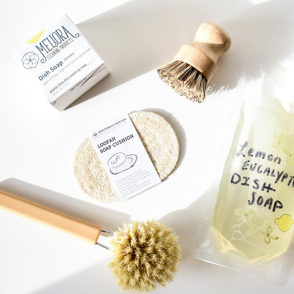 Top-down view of eco-friendly dishwashing items: Meliora dish soap box, compostable round loofah soap cushion, two Redecker Dish Brushes with Removable Head (natural bristles, wooden handles), and a pouch labeled lemon eucalyptus dish soap.