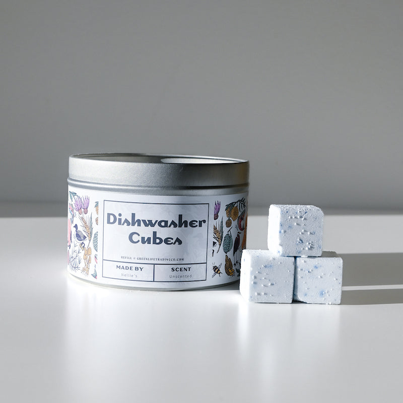 A silver tin of Nellies Dishwasher Cube with a floral label sits on a white surface beside three white, speckled dishwasher cubes.