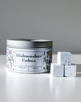 A silver tin of Nellies Dishwasher Cube with a floral label sits on a white surface beside three white, speckled dishwasher cubes.