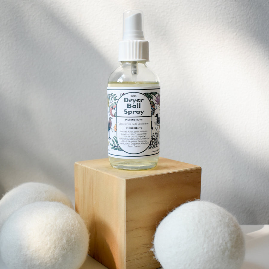 A clear spray bottle of Shade Tree Naturals Linen & Dryer Ball Mist sits on a wooden block, surrounded by three white wool dryer balls, with a soft, light-textured background.