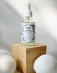 A clear spray bottle of Shade Tree Naturals Linen & Dryer Ball Mist sits on a wooden block, surrounded by three white wool dryer balls, with a soft, light-textured background.