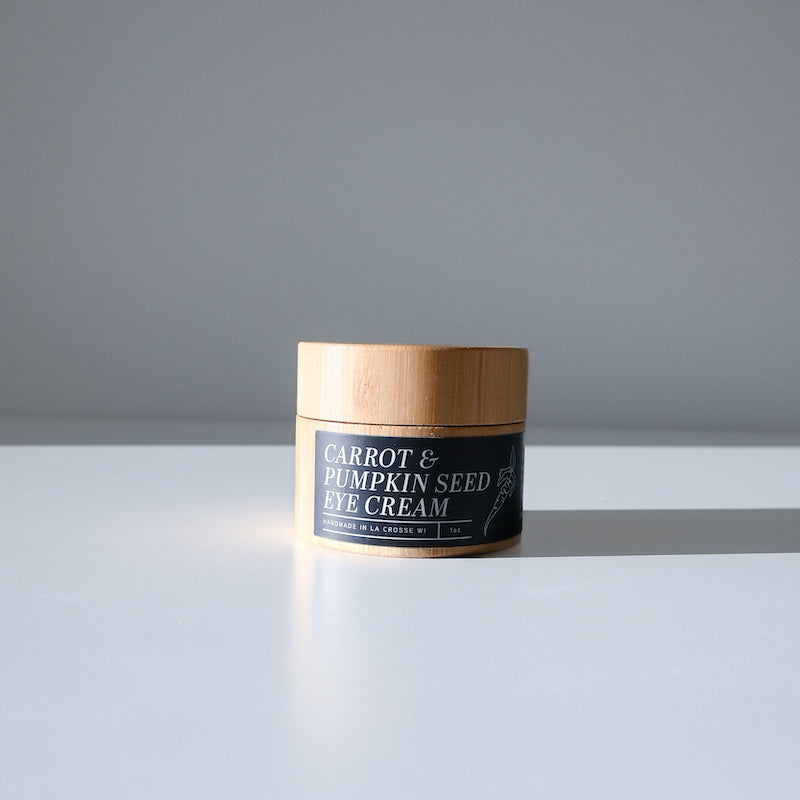 A small jar of Larsons General Carrot and Pumpkin Seed Eye Cream with a wooden lid rests on a white surface, casting a gentle shadow on a gray background and highlighting its nourishing, moisturizing formula.