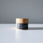 A small jar of Larsons General Carrot and Pumpkin Seed Eye Cream with a wooden lid rests on a white surface, casting a gentle shadow on a gray background and highlighting its nourishing, moisturizing formula.