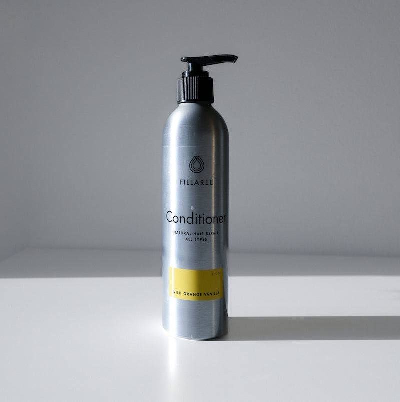 A silver pump bottle of Fillaree Conditioner by Fillaree rests on a white surface with a gray background. The sulfate-free, hydrating formula suits all hair types and features a wild orange vanilla scent.