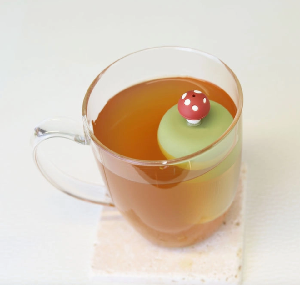 A glass mug from Teany Tiny Kitchen features the Floating Tea Infuser—a green silicone lid with a mushroom-shaped handle—ideal for keeping your tea warm and optimizing steeping.