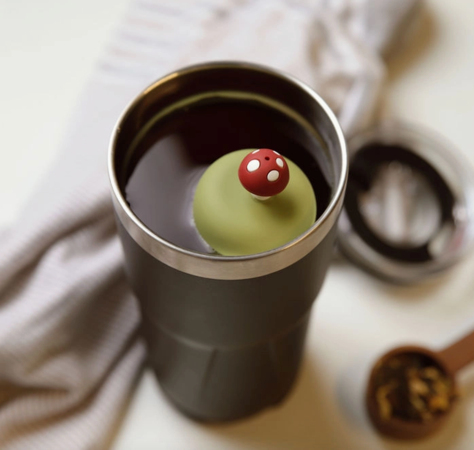 The Teany Tiny Kitchen Floating Tea Infuser sits in a tumbler of dark tea, topped with a green lid featuring a red, white-spotted mushroom handle. A measuring spoon and textured cloth suggest tea is being steeped right now.