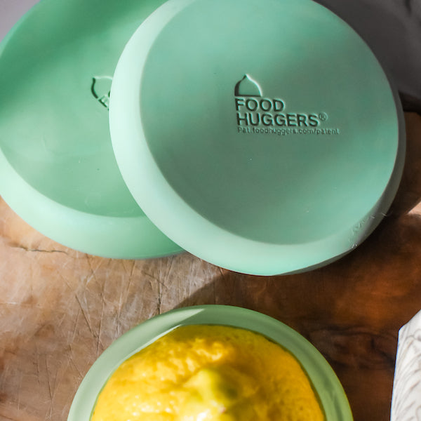 A lemon half sits on a wooden surface, covered with a green Food Huggers silicone lid. Two more lids are nearby, each embossed with the Food Huggers logo—an easy way to reduce food waste in your kitchen.