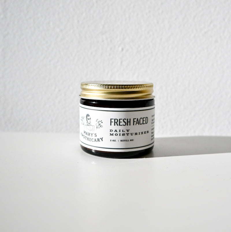A small jar of Fresh Faced Daily Moisturizer from Larsons General rests on a white surface near a light gray textured wall, its lightweight formula offering smooth skin as sunlight creates a soft shadow next to it.