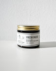 A small jar of Fresh Faced Daily Moisturizer from Larsons General rests on a white surface near a light gray textured wall, its lightweight formula offering smooth skin as sunlight creates a soft shadow next to it.