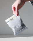 A hand holds a white paper bag labeled tea against a plain light gray background.
