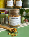 A refillable Jar of Golden Milk tea the jar has a cork lid and an attachable bamboo spoon. the spoon is in front full of loose golden milk tea.