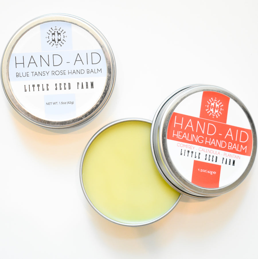 Two metal tins of Little Seed Farm Healing Hand Balm, one open to reveal the yellow balm inside. Labels indicate organic ingredients and feature the product name: Healing Hand Balm.