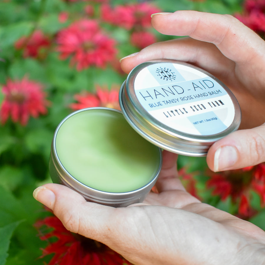 A person opens a small tin of Little Seed Farm Healing Hand Balm, green in color, with a blurred background of vibrant red garden flowers.
