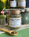 A refillable jar of happy tummy tea. The jar has a cork lid and an attachable bamboo spoon. The spoon is in front of the jar with tea in it.
