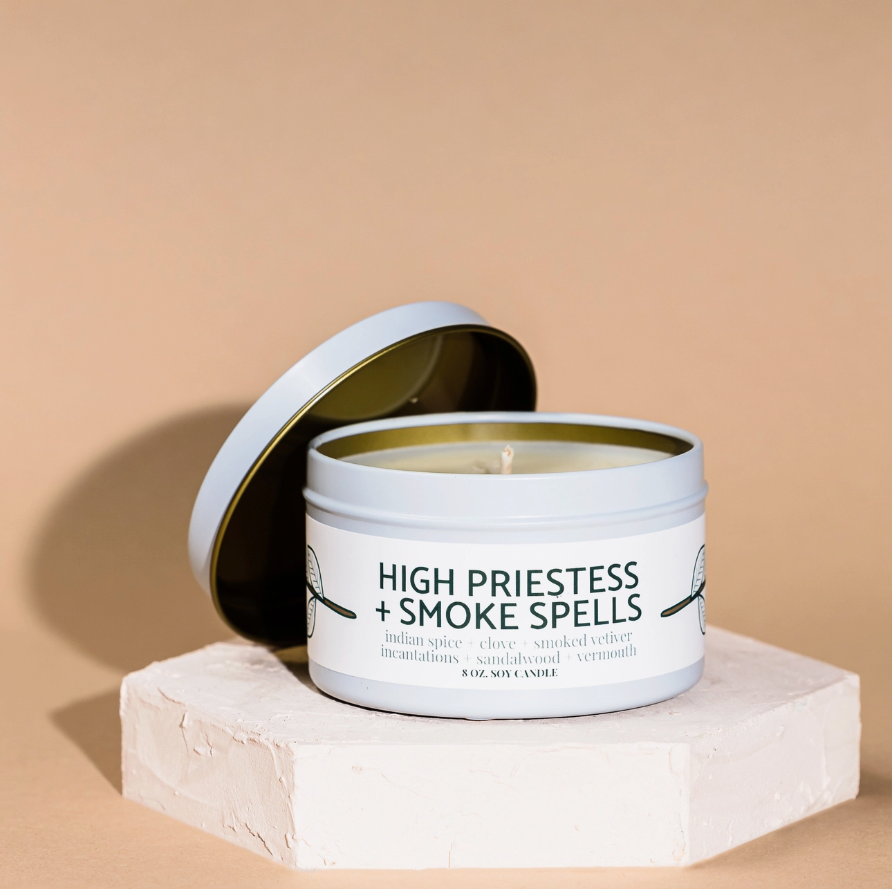 A Modern Maker Provisions Soy Candle by Modern Makers sits on a textured white platform with its lid to the side. This hand-poured soy candle offers a long-lasting fragrance against a neutral beige background.