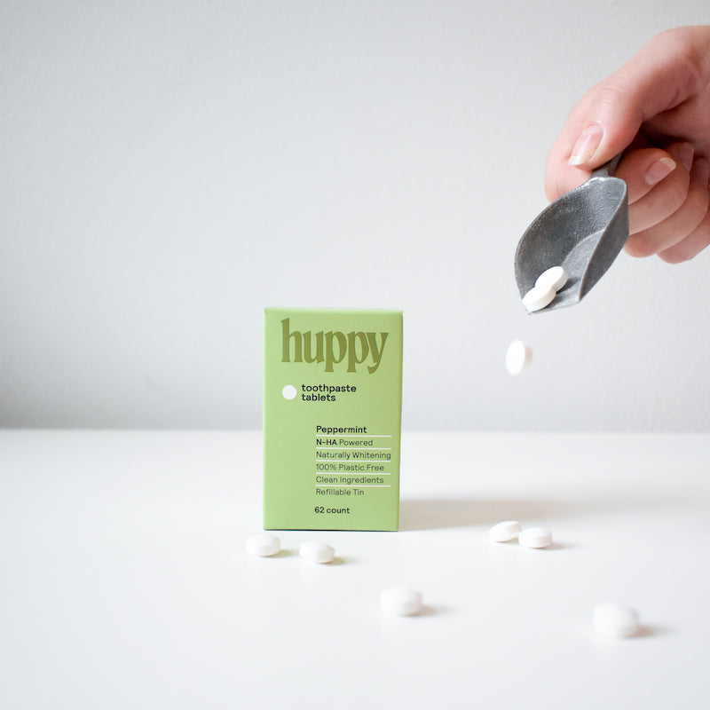 A hand pours white Huppy Foaming Tooth Tabs from a metal scoop next to a green Huppy box, with several tabs scattered on a white surface—a refreshing toothpaste alternative.