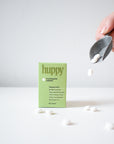 A hand pours white Huppy Foaming Tooth Tabs from a metal scoop next to a green Huppy box, with several tabs scattered on a white surface—a refreshing toothpaste alternative.