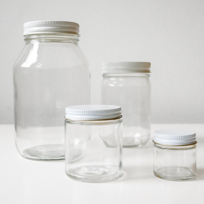 Four empty Container Glass Jars with aluminum caps in different sizes are displayed on a white surface, ideal for storing your DIY creations against a light background.