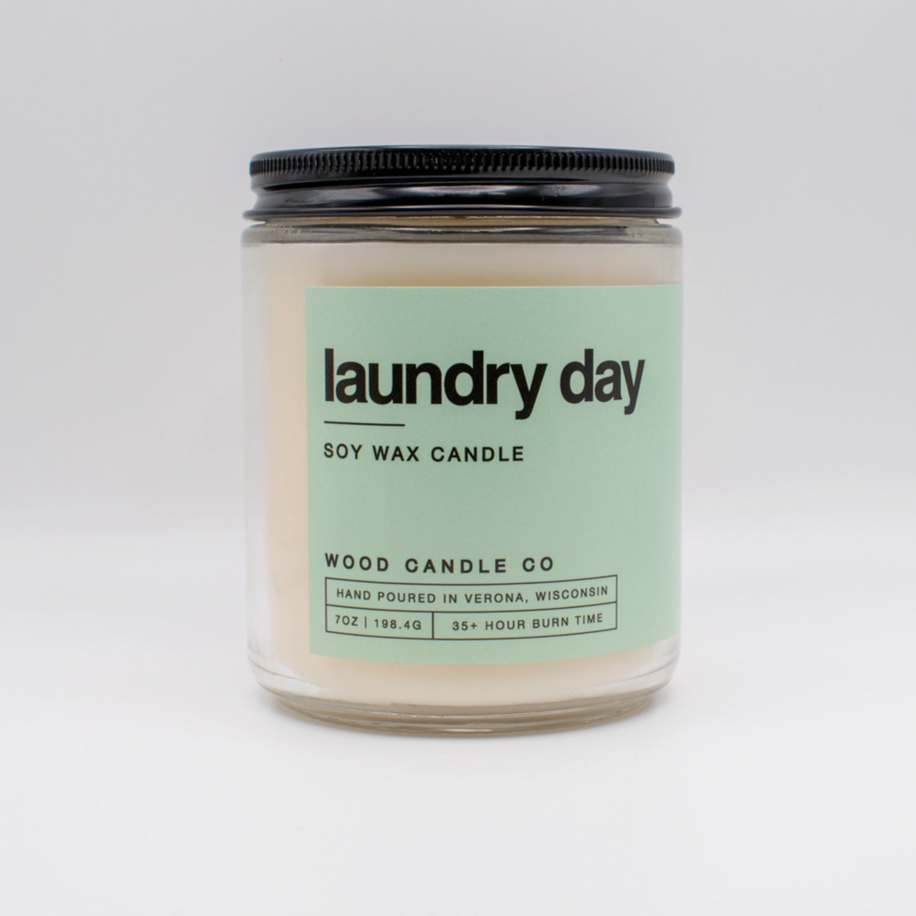 A clear glass jar candle with a black lid, labeled “laundry day” in bold, features a mint-green label for Wood Candle Co. Soy Candle. Hand-poured in Verona, Wisconsin, it offers long-lasting fragrance and a 35+ hour burn time.