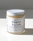 A clear jar with a white lid labeled Shade Tree Naturals Sugar Scrub Lavender Lemongrass sits on a white surface. This emulsified scrub gently exfoliates—apply a small amount to damp skin and massage in the shower.