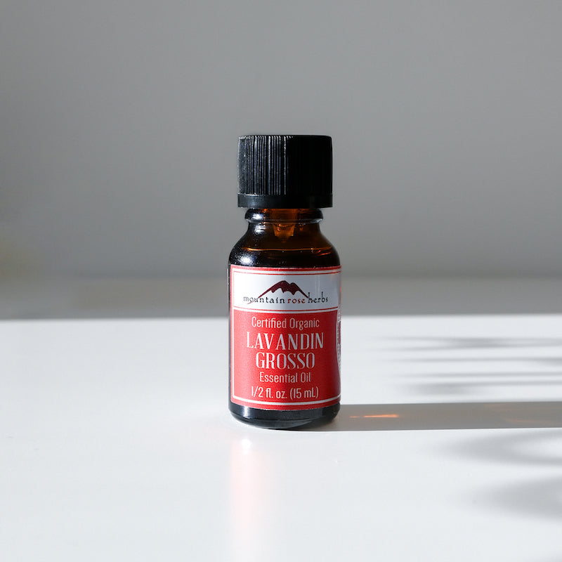 A 0.5 fl oz (15 mL) amber bottle of Mountain Rose Herbs Lavender Essential Oil rests on a white surface in bright light, casting a shadow. This certified organic oil offers a floral aroma ideal for aromatherapy.