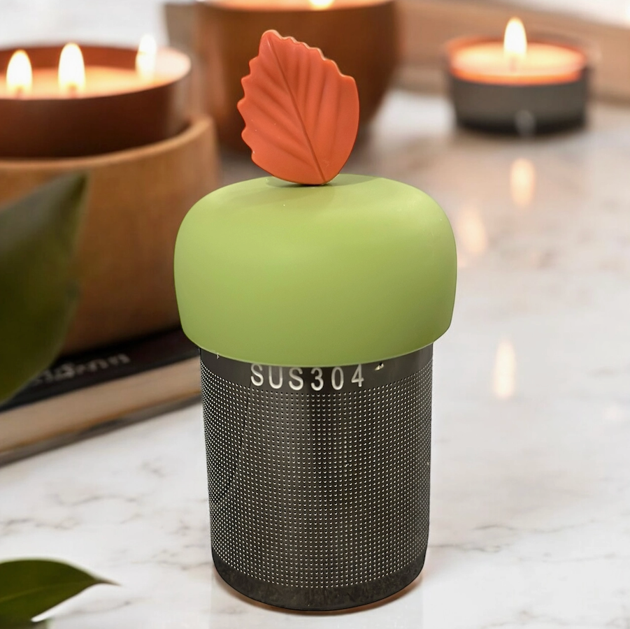 The Teany Tiny Kitchen Floating Tea Infuser features a stainless steel body, green silicone top, and orange leaf-shaped handle, shown on a marble countertop with softly blurred candles and a plant in the background.