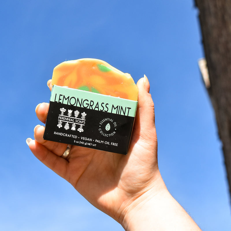 A hand holds up a vibrant Bar Soap by Perennial Soaps against a clear blue sky, showcasing its Wisconsin-made, handcrafted, vegan, and palm oil free qualities.