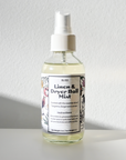 A clear spray bottle of Shade Tree Naturals Linen & Dryer Ball Mist, with floral designs and instructions to add distilled water and essential oils, rests on a white surface in bright natural light against a simple white background.
