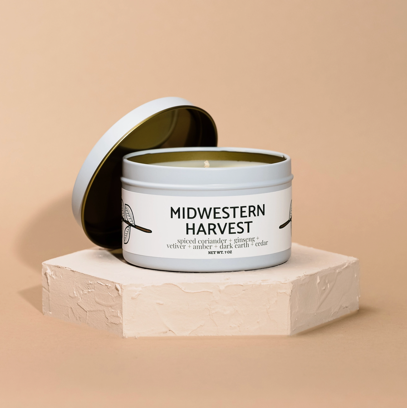 A white tin Modern Maker Provisions Soy Candle by Modern Makers, hand-poured in small batches, sits on a textured beige platform. Its label highlights spiced coriander, vetiver, and cedar for a long-lasting scent.