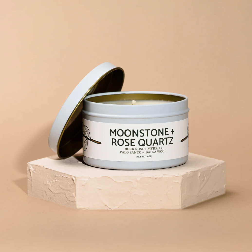 A white metal tin Modern Maker Provisions Soy Candle by Modern Makers, with its lid nearby, sits on a light textured pedestal against a beige background—an elegant hand-poured soy candle designed for lasting fragrance.