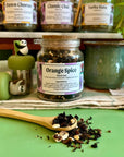 A refillable jar of orange spice loose leaf tea. The jar has a cork lid and an attachable bamboo spoon. The spoon is in front full of orange spice tea. Behind the jar is a shelf full of tea jars.