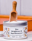 An open metal tin of Meliora Oxy Brightener & Spot Remover with a floral design displays eco-friendly white powder inside, topped with a small wooden scoop resting upright.