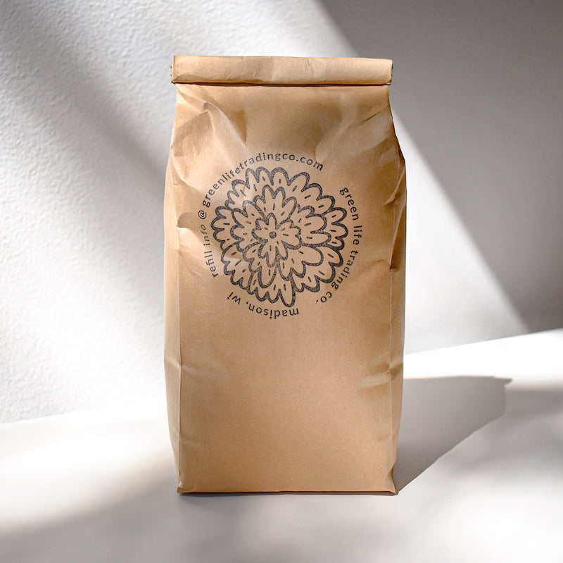 A brown paper bag labeled Jasmine Green Tea from Mountain Rose Herbs stands upright on a white surface, displaying a circular floral logo and text, with sunlight casting nearby shadows.