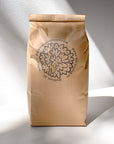 A brown paper bag labeled Jasmine Green Tea from Mountain Rose Herbs stands upright on a white surface, displaying a circular floral logo and text, with sunlight casting nearby shadows.