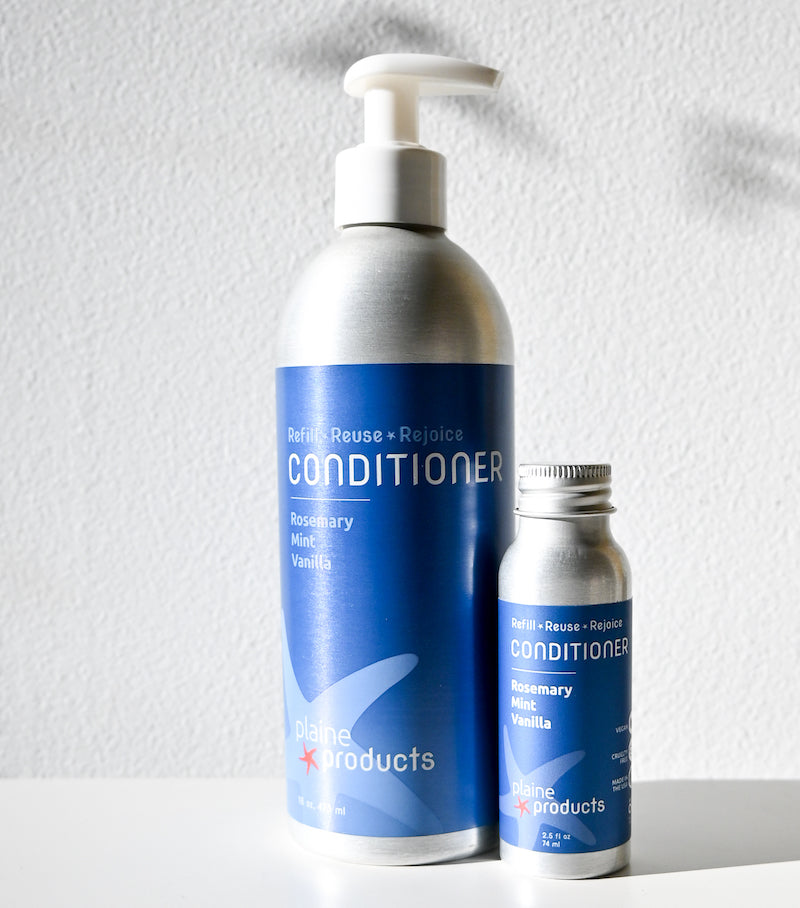 Two Plaine Products Conditioner bottles by Plaine Products—one large with a pump, one small refillable—sit on a white surface. The salon-quality, sulfate-free formula features rosemary, mint, and vanilla scents and has blue labels on the aluminum containers.