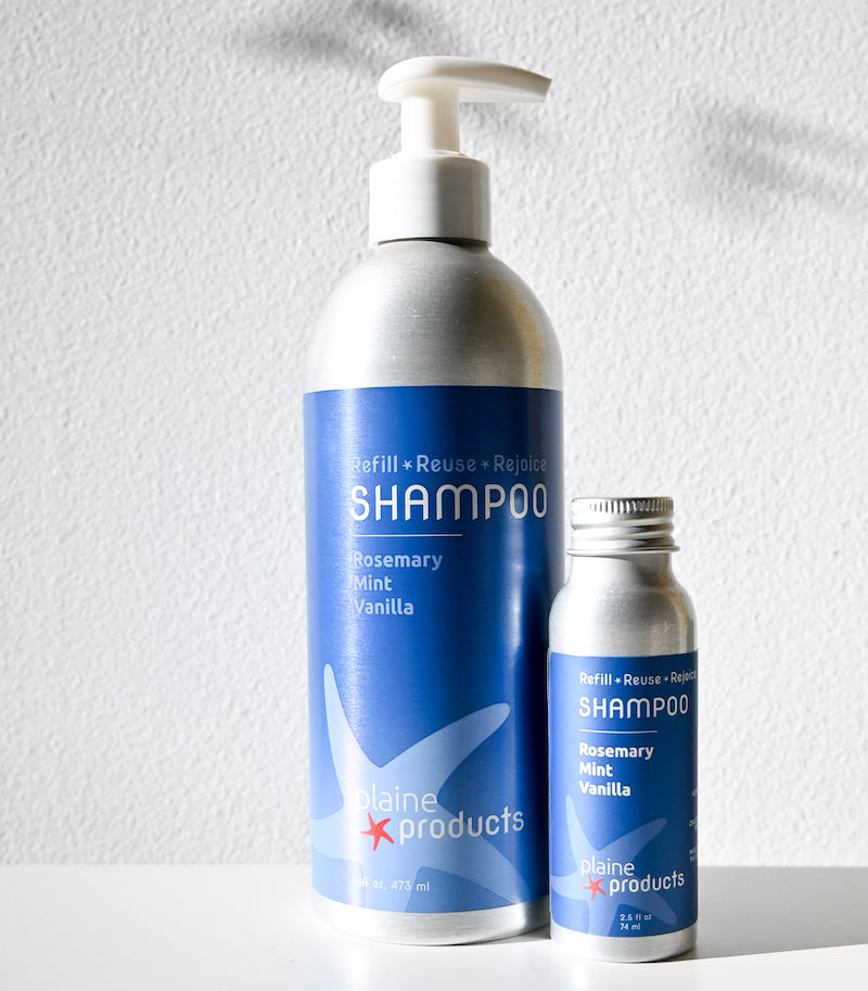 Two silver bottles of Plaine Products Shampoo—one large with a pump, one small with a screw cap—feature blue labels with a starfish design. This vegan, sulfate-free shampoo is scented Rosemary Mint Vanilla and comes in refillable packaging.